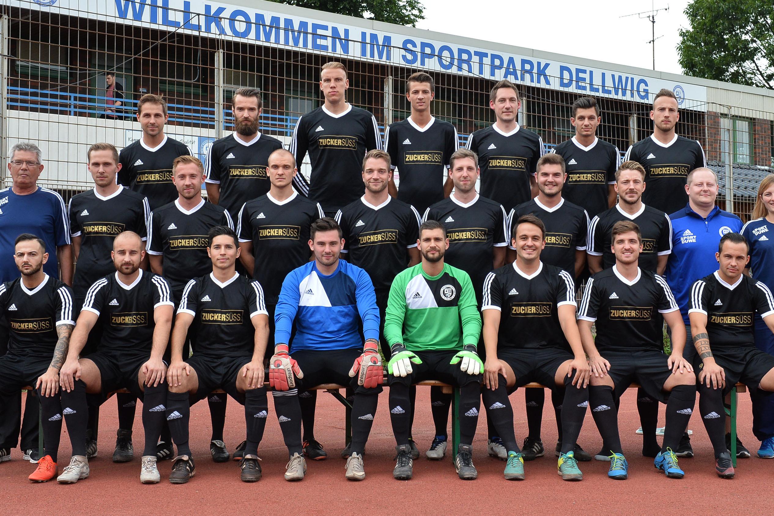 Gruppenfoto einer Fu&szlig;ballmannschaft in schwarz-wei&szlig;er Sportkleidung im Sportpark Dellwig vor einem Begr&uuml;&szlig;ungsschild.