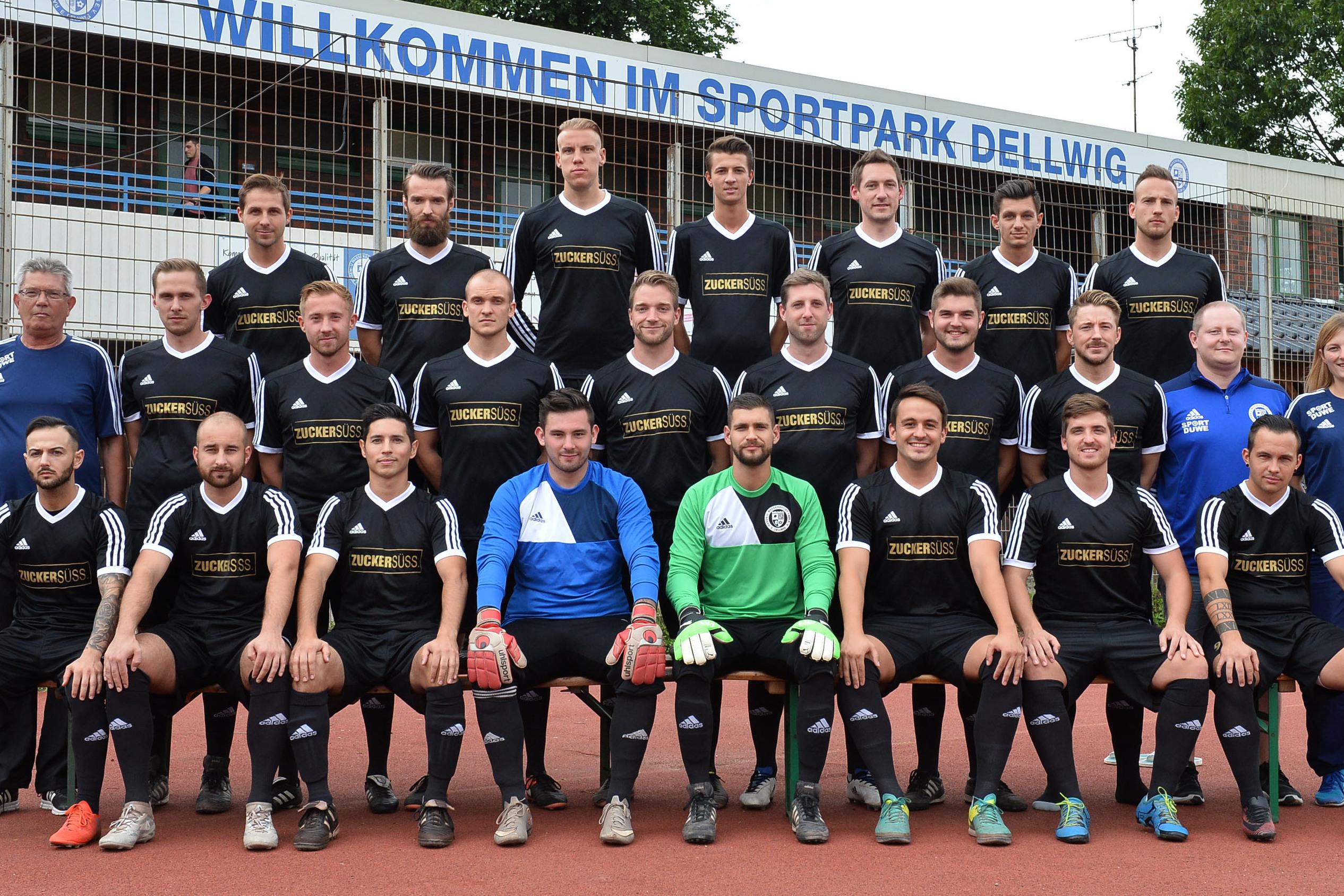 Gruppenfoto einer Fu&szlig;ballmannschaft in schwarz-wei&szlig;er Sportkleidung im Sportpark Dellwig vor einem Begr&uuml;&szlig;ungsschild.