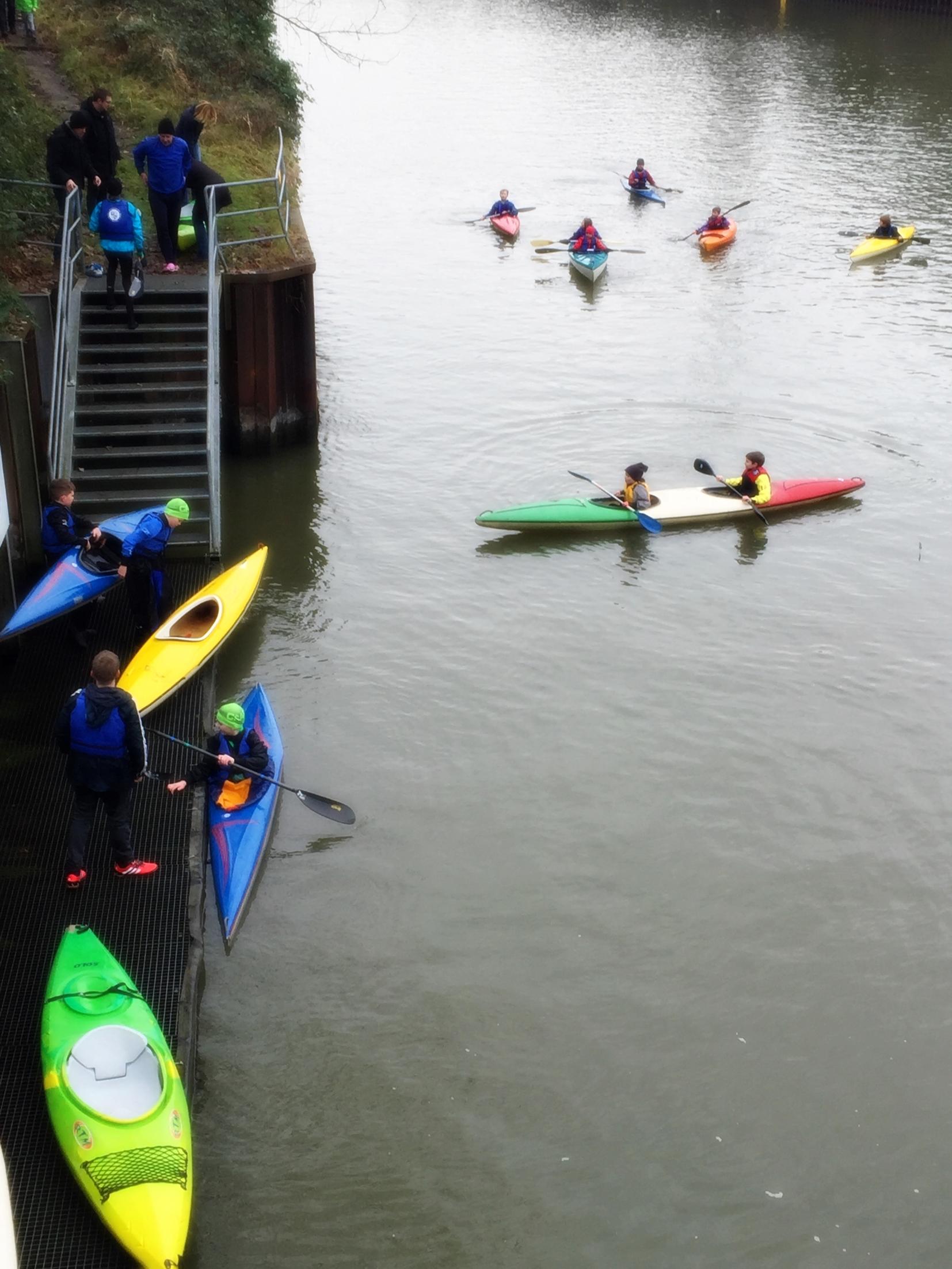 Gruppierung von Paddlern in Kajaks auf einem Gew&auml;sser, einige steigen &uuml;ber Treppen ein, andere paddeln.