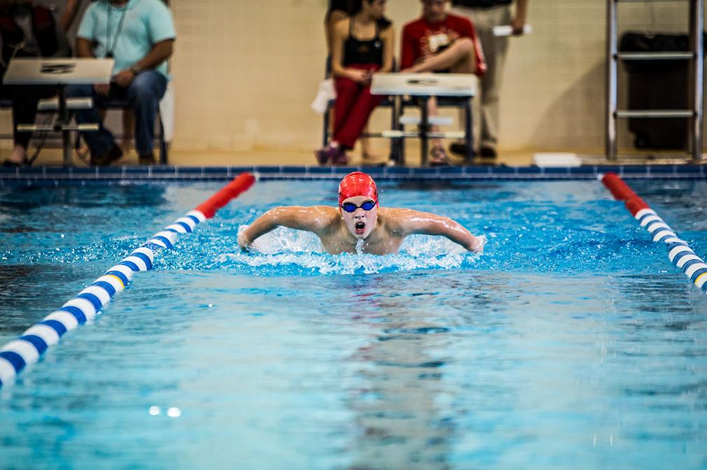 Schwimmer in roter Badekappe und Brille Brustschwimmen im blauen Schwimmbecken, Zuschauer im Hintergrund.