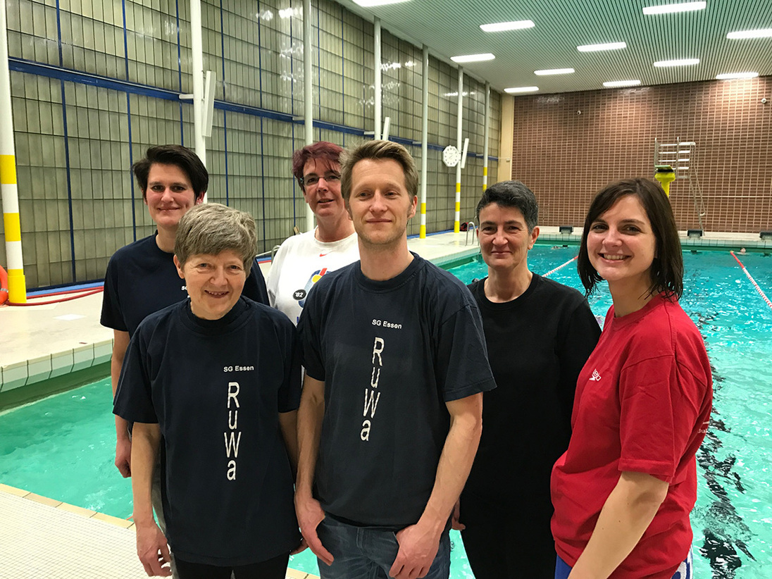 Sechs Personen in Schwimmkleidung stehen l&auml;chelnd neben einem Schwimmbecken in einer Sporthalle.