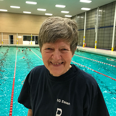 L&auml;chelnde Frau mit grauen Haaren in einem Schwimmbad, im Hintergrund schwimmen mehrere Sportler.