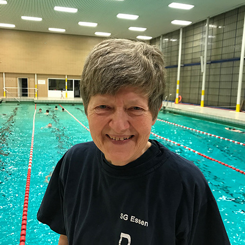 L&auml;chelnde Frau mit grauen Haaren in einem Schwimmbad, im Hintergrund schwimmen mehrere Sportler.