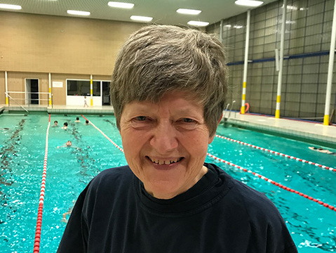 L&auml;chelnde Frau mit grauen Haaren in einem Schwimmbad, im Hintergrund schwimmen mehrere Sportler.