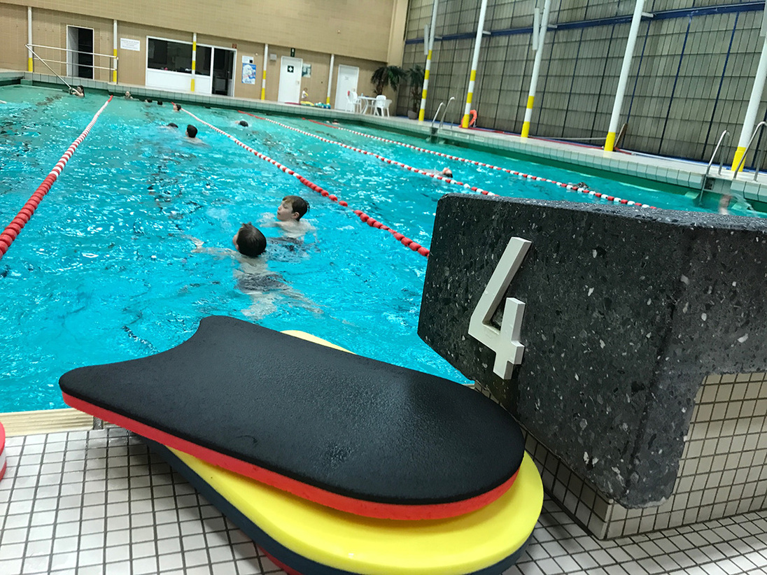 Nahaufnahme von Schwimmbrett und Startblock mit der Nummer 4 an einem geschwungenen Schwimmbecken mit schwimmenden Personen.