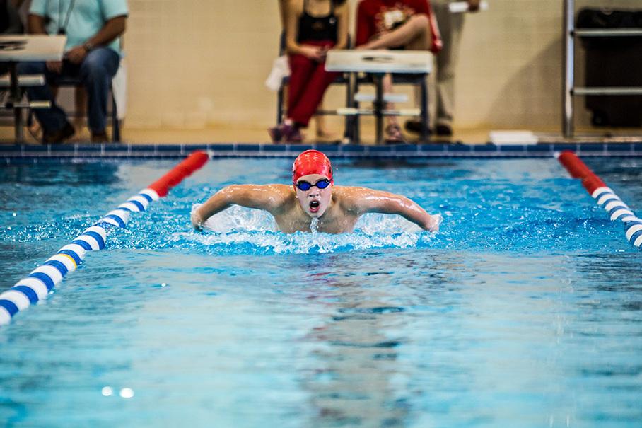 Schwimmer in roter Badekappe und Brille Brustschwimmen im blauen Schwimmbecken, Zuschauer im Hintergrund.
