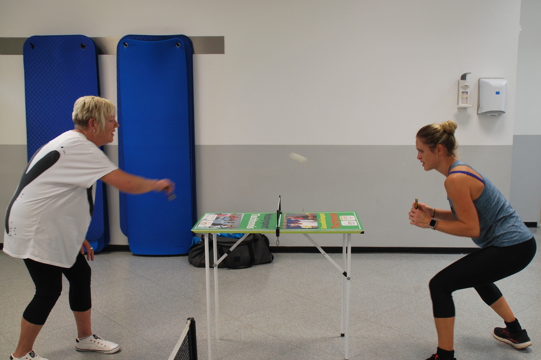 Zwei Frauen spielen Tischtennis an einem kleinen Tisch in einem modernen Trainingsraum.