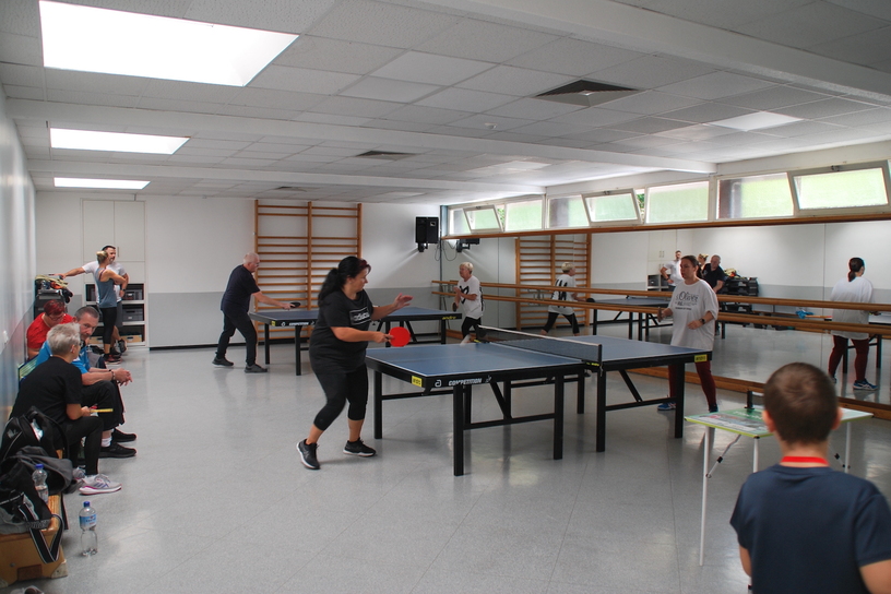 Tischtennis-Spieler trainieren in einem hellen Raum mit mehreren Tischtennisplatten und Zuschauern am Rand.