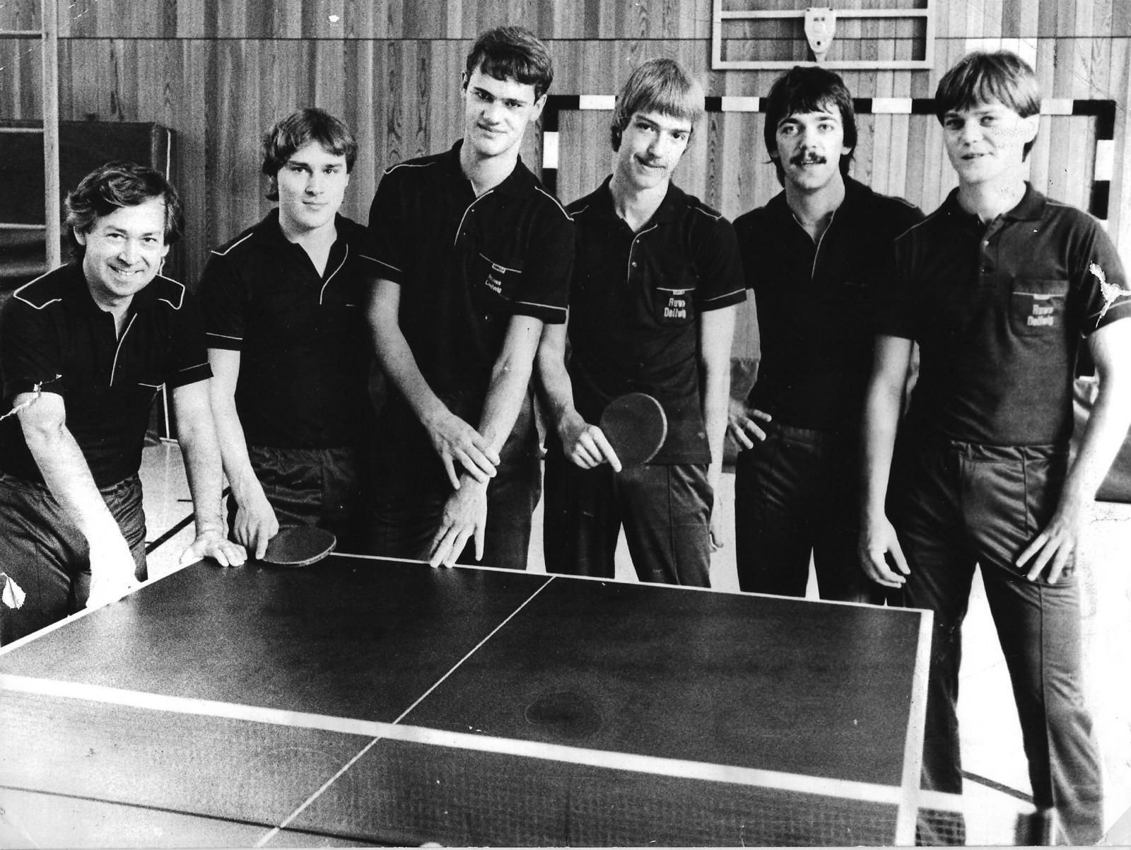 Sechs Tischtennisspieler in dunklen Polohemden stehen um einen Tischtennistisch in einer Sporthalle.