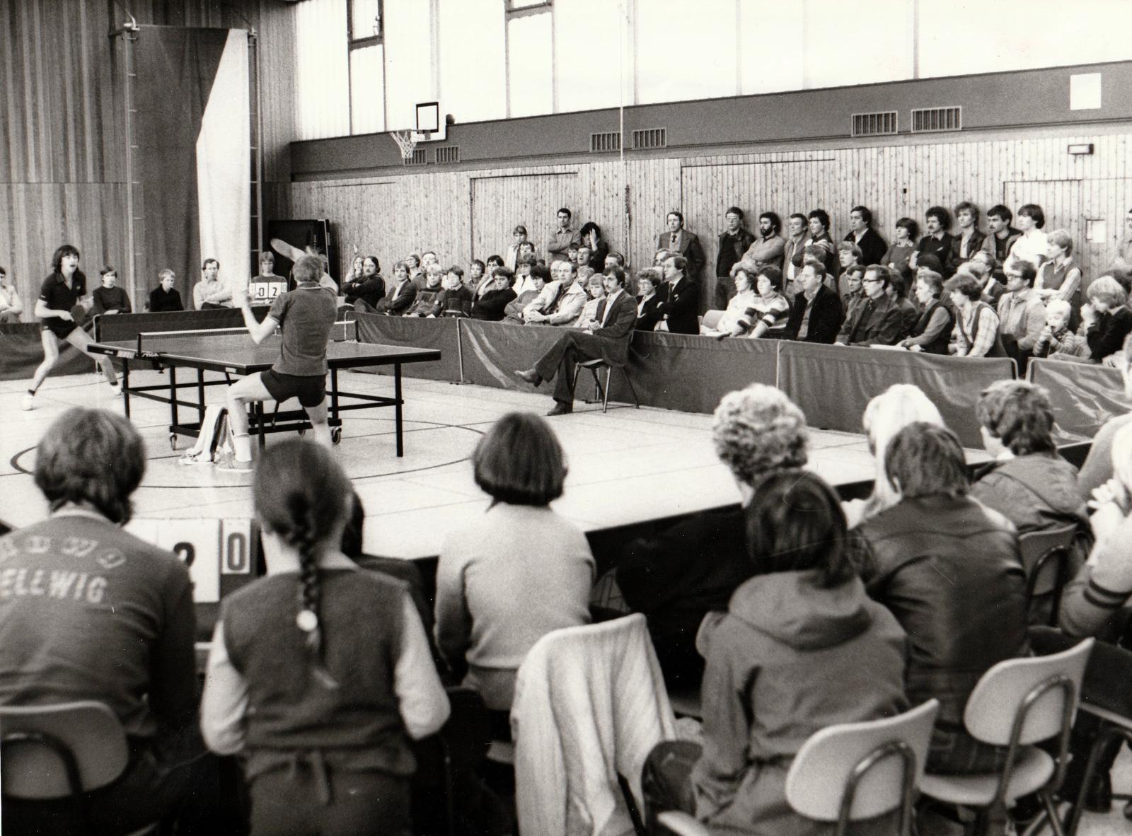 Tischtennisturnier: Spielerinnen am Tisch, Publikum auf B&auml;nken beobachtet das Geschehen in einer Sporthalle.