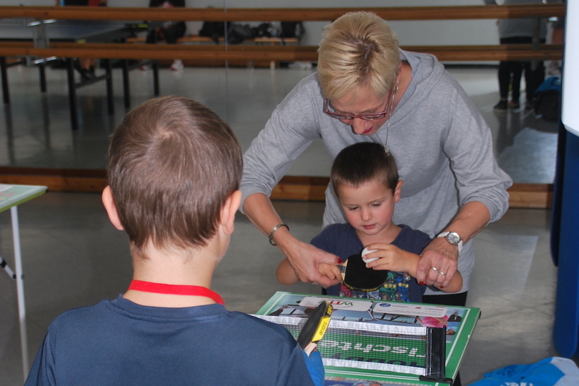 Kinder stehend an einem Tisch, w&auml;hrend eine Frau ihnen beim Basteln an einem Tischtennis-Tisch hilft.