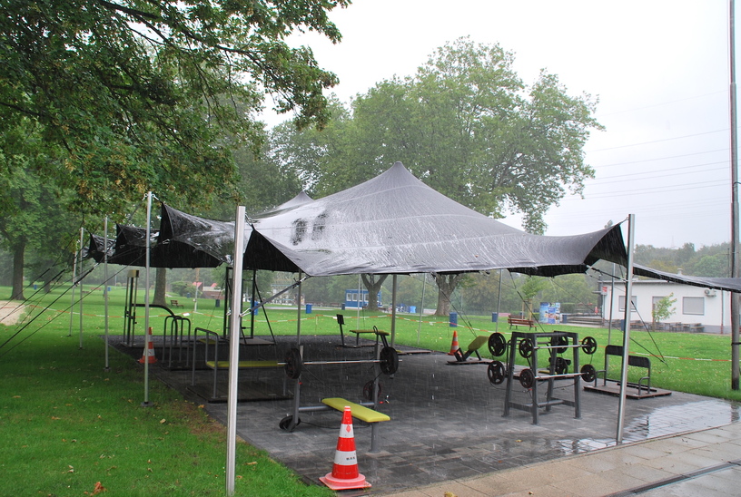 Zelt mit Gewichten und Fitnessger&auml;ten auf einem grassierten Platz bei Regenwetter, umgeben von B&auml;umen.