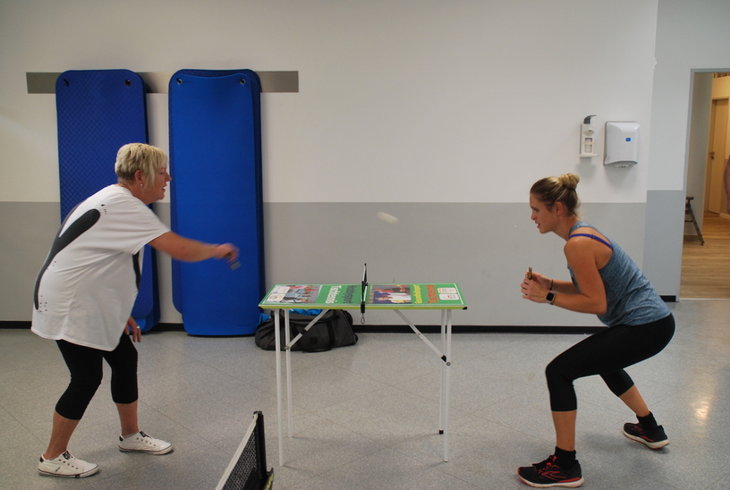 Zwei Frauen spielen Tischtennis an einem kleinen Tisch in einem modernen Trainingsraum.