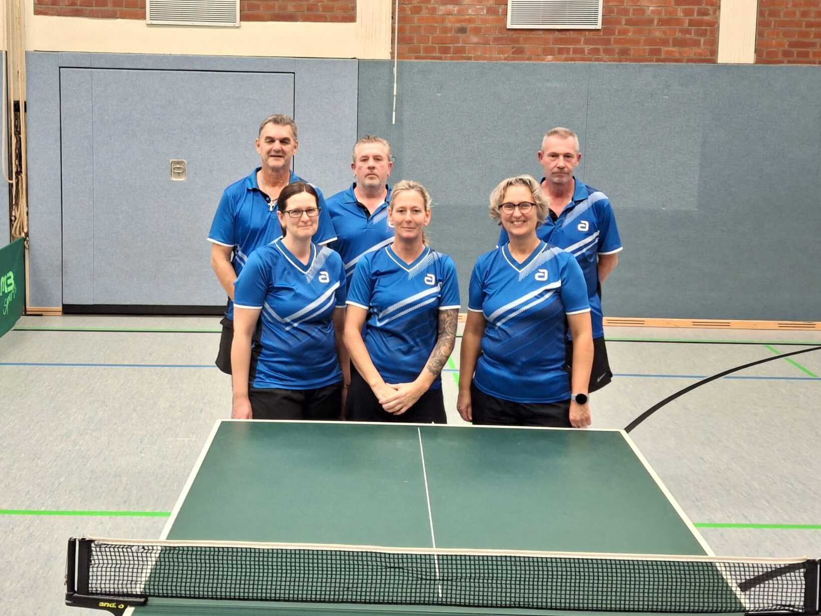 Sechs Tischtennisspieler in blauen Trikots stehen hinter einem Tischtennistisch in einer Sporthalle.