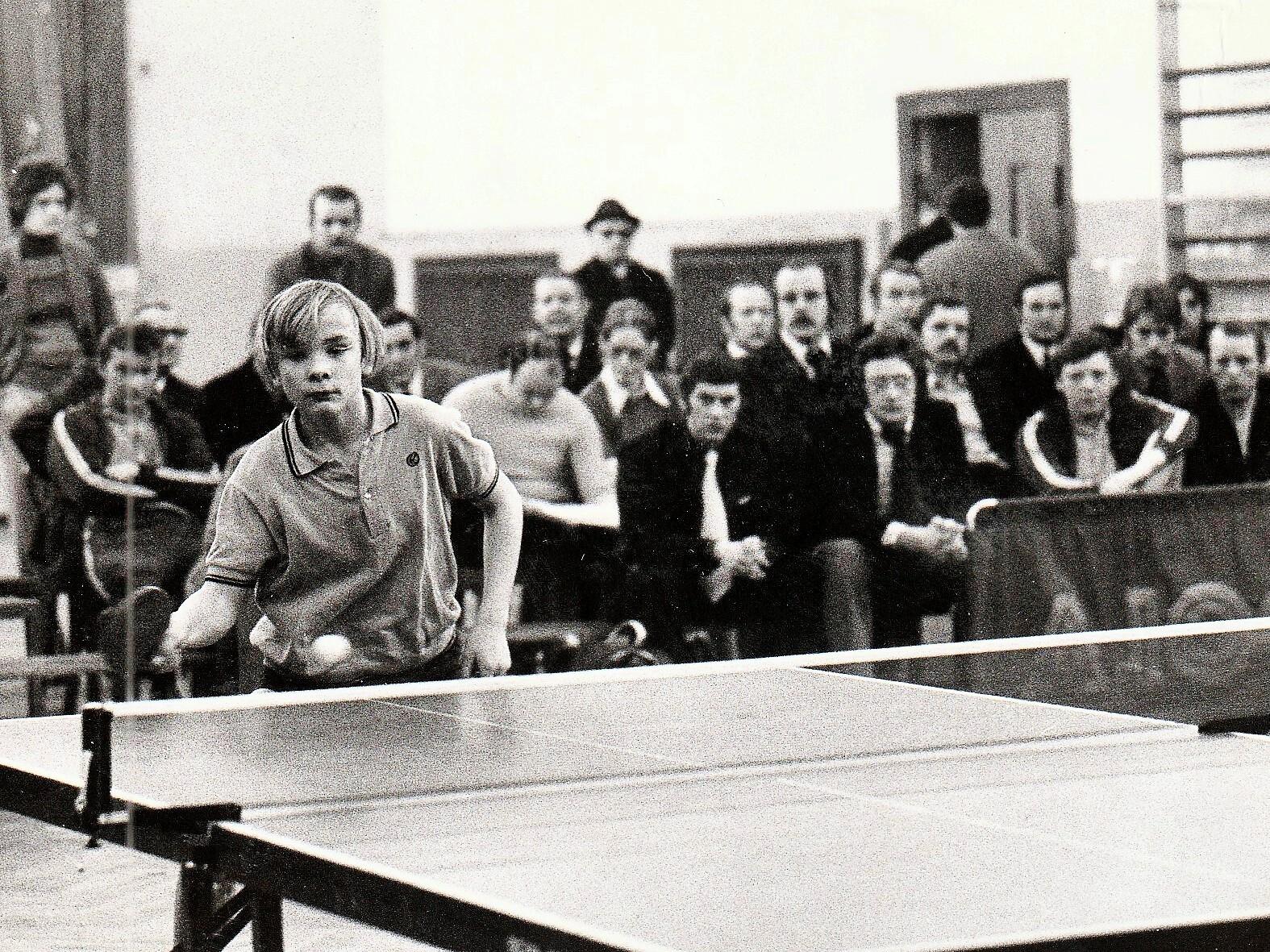 Junge spielt Tischtennis, w&auml;hrend Zuschauer im Hintergrund auf einer Sportveranstaltung zuschauen.
