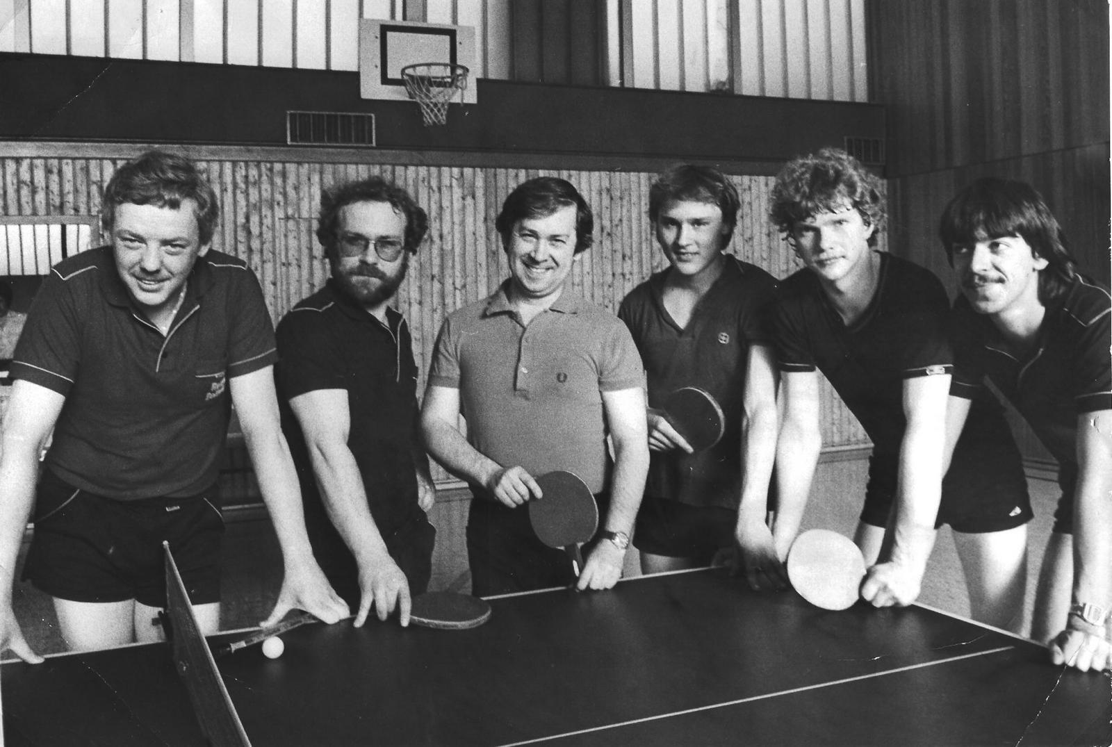 Sechs M&auml;nner posieren mit Tischtennisschl&auml;gern vor einer Tischtennisplatte in einer Sporthalle.