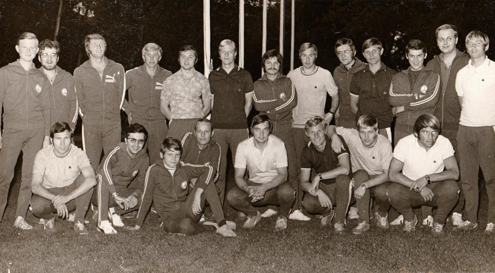 Gruppenfoto von 16 M&auml;nnern in Sportkleidung, die auf einer Wiese posieren, umgeben von B&auml;umen und Lichtmasten.