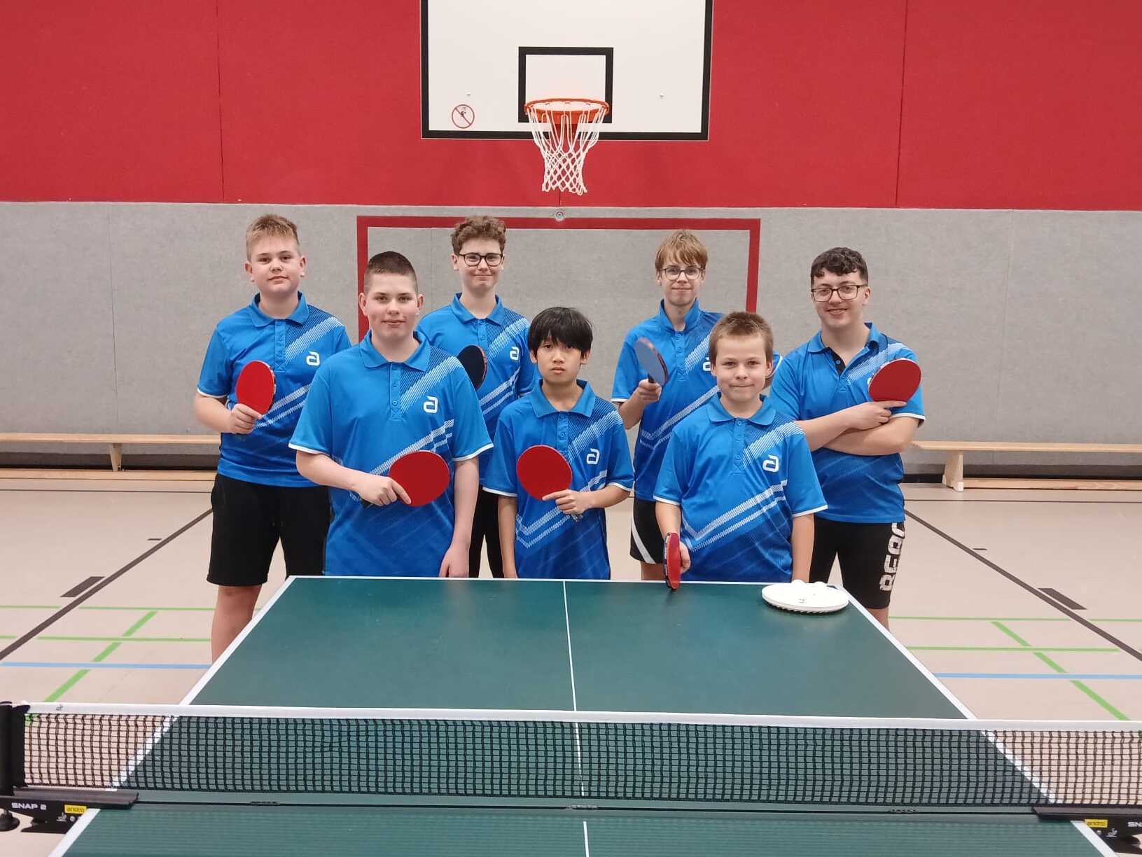 Gruppe von sieben Tischtennisspielern in blauen Trikots steht lächelnd vor einem Tischtennistisch.