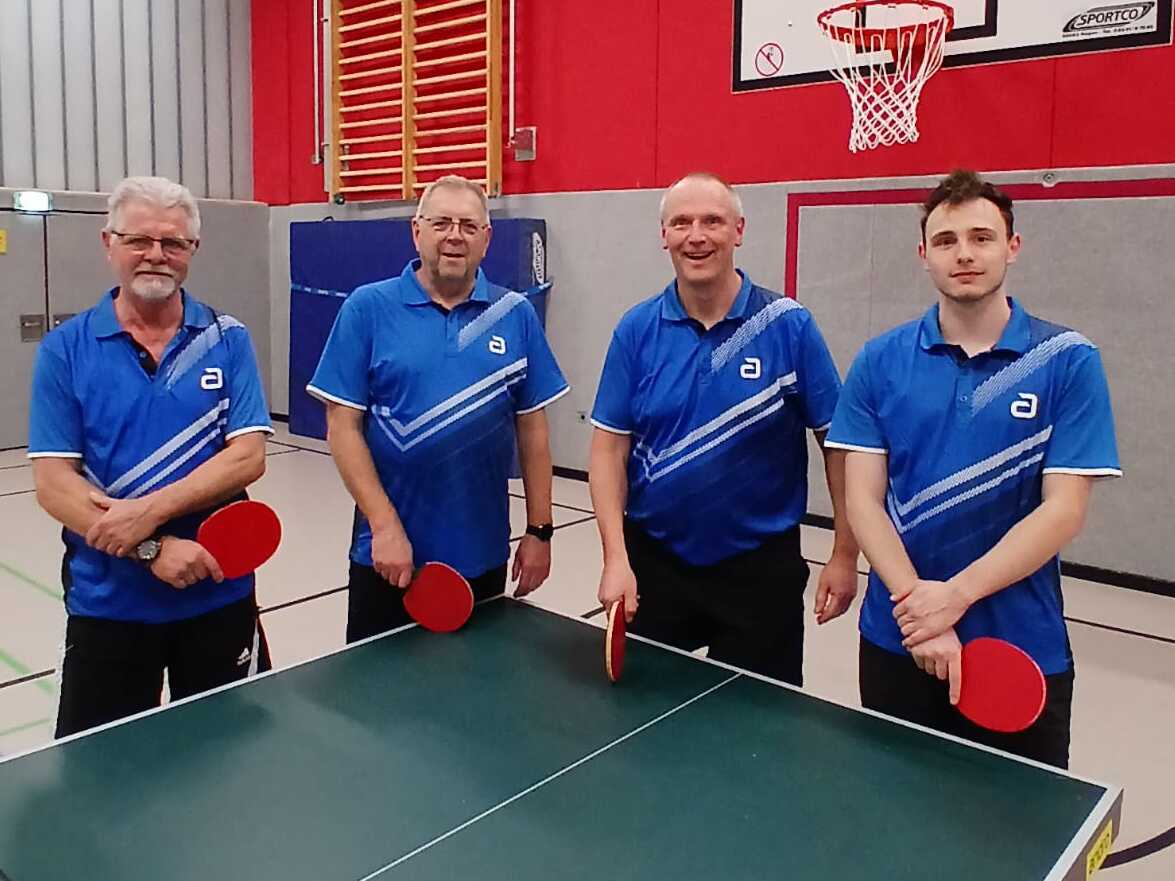 Vier M&auml;nner in blauen Sporthemden stehen an einem Tischtennistisch mit Schl&auml;gern. Im Hintergrund ist ein Basketballkorb sichtbar.