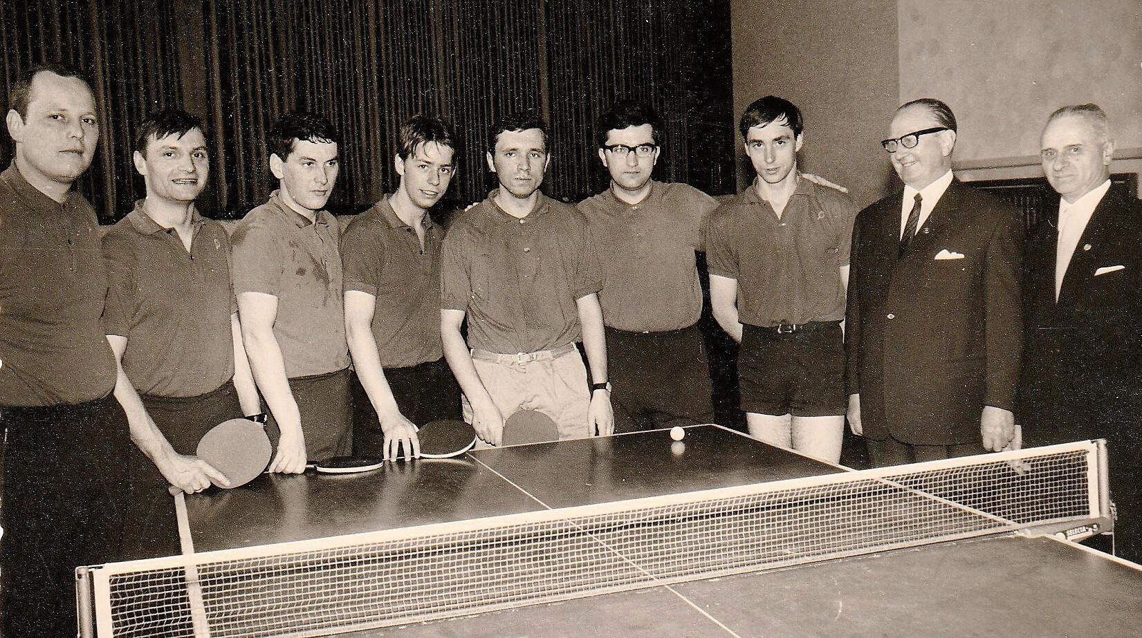 Gruppe von sieben Tischtennisspielern und zwei Offiziellen neben einem Tischtennistisch mit Netz und Ball.