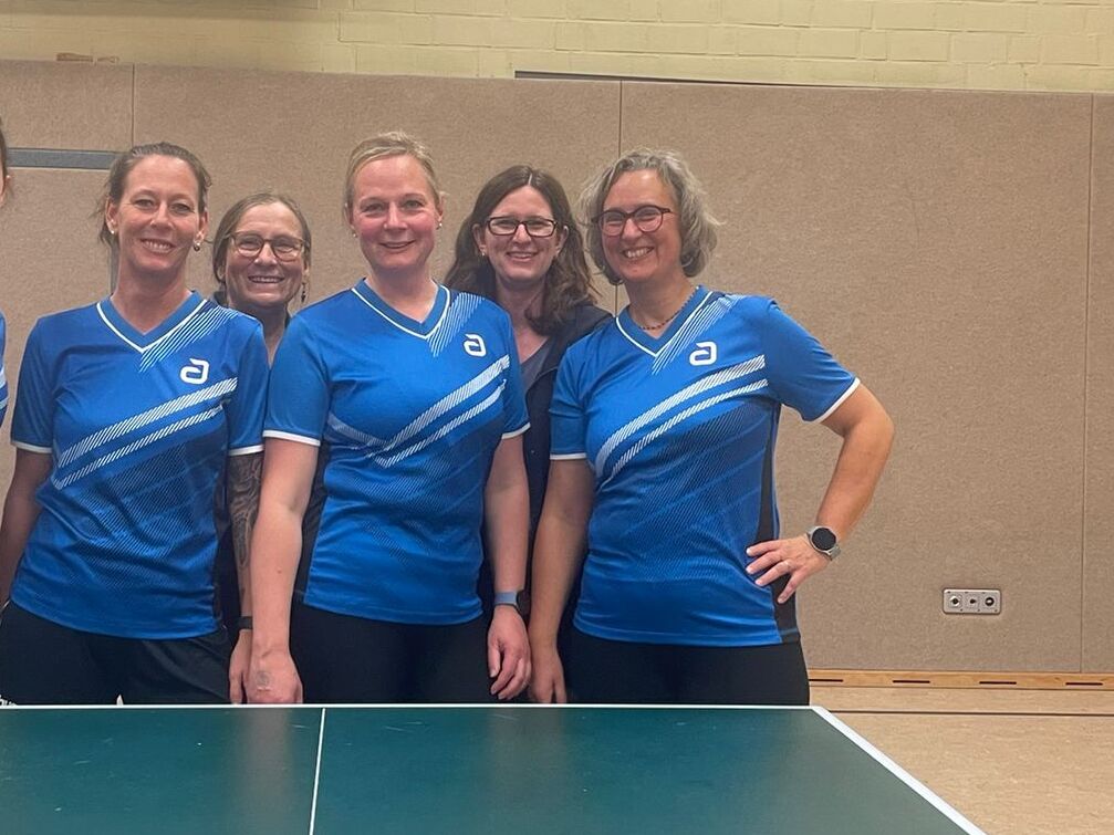 Sechs Frauen in blauen T-Shirts posieren l&auml;chelnd vor einem Tischtennistisch in einer Sporthalle.