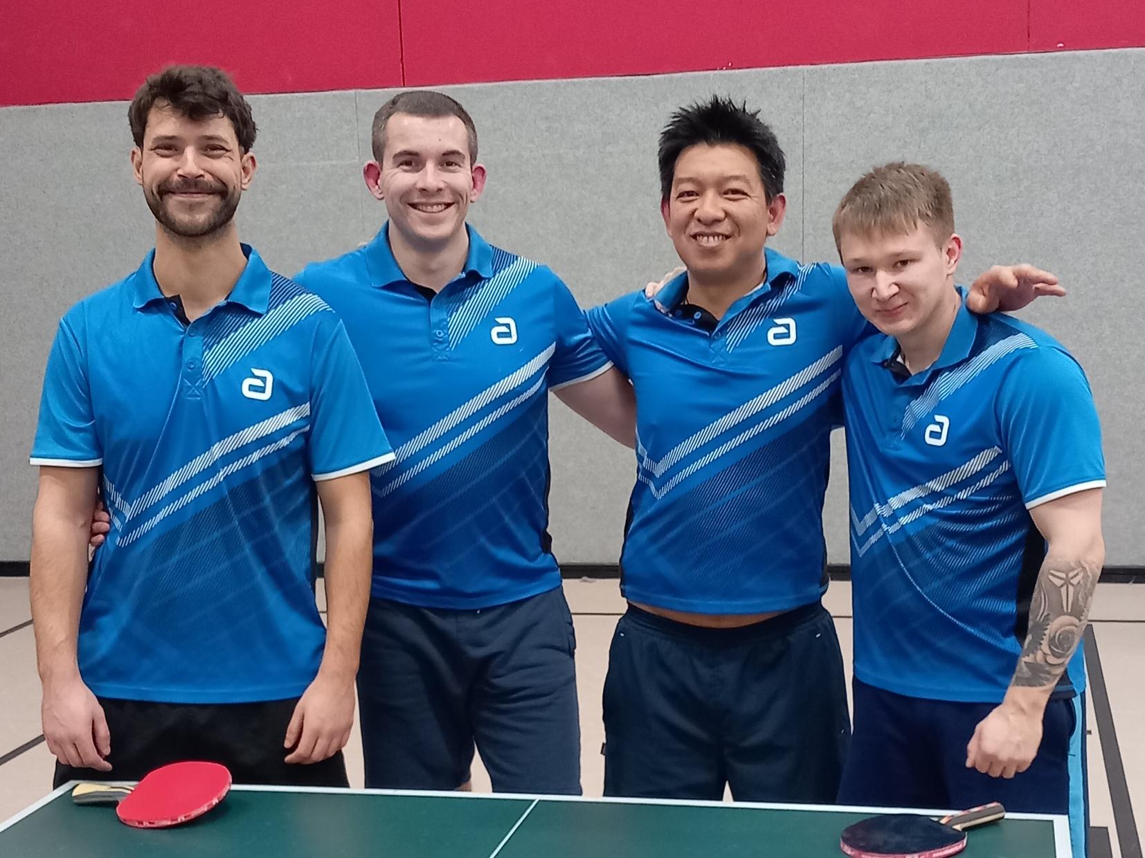 Vier Tischtennisspieler in blauen Trikots stehen l&auml;chelnd vor einem Tischtennistisch mit Schl&auml;gern und B&auml;llen.