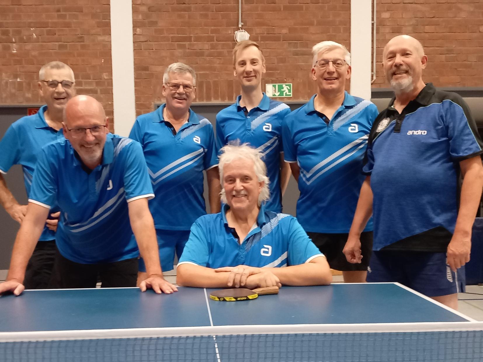 Gruppe von sieben männlichen und weiblichen Tischtennisspielern in blauen Trikots, lächelnd hinter einem Tischtennistisch.