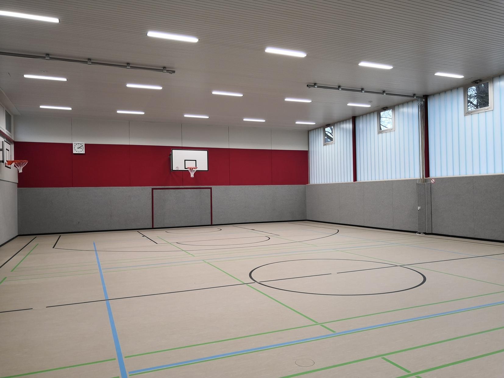Leere Turnhalle mit grauem Boden, roten Wänden und zwei Basketballkörben an einer Wand. Tageslicht fällt durch Fenster.