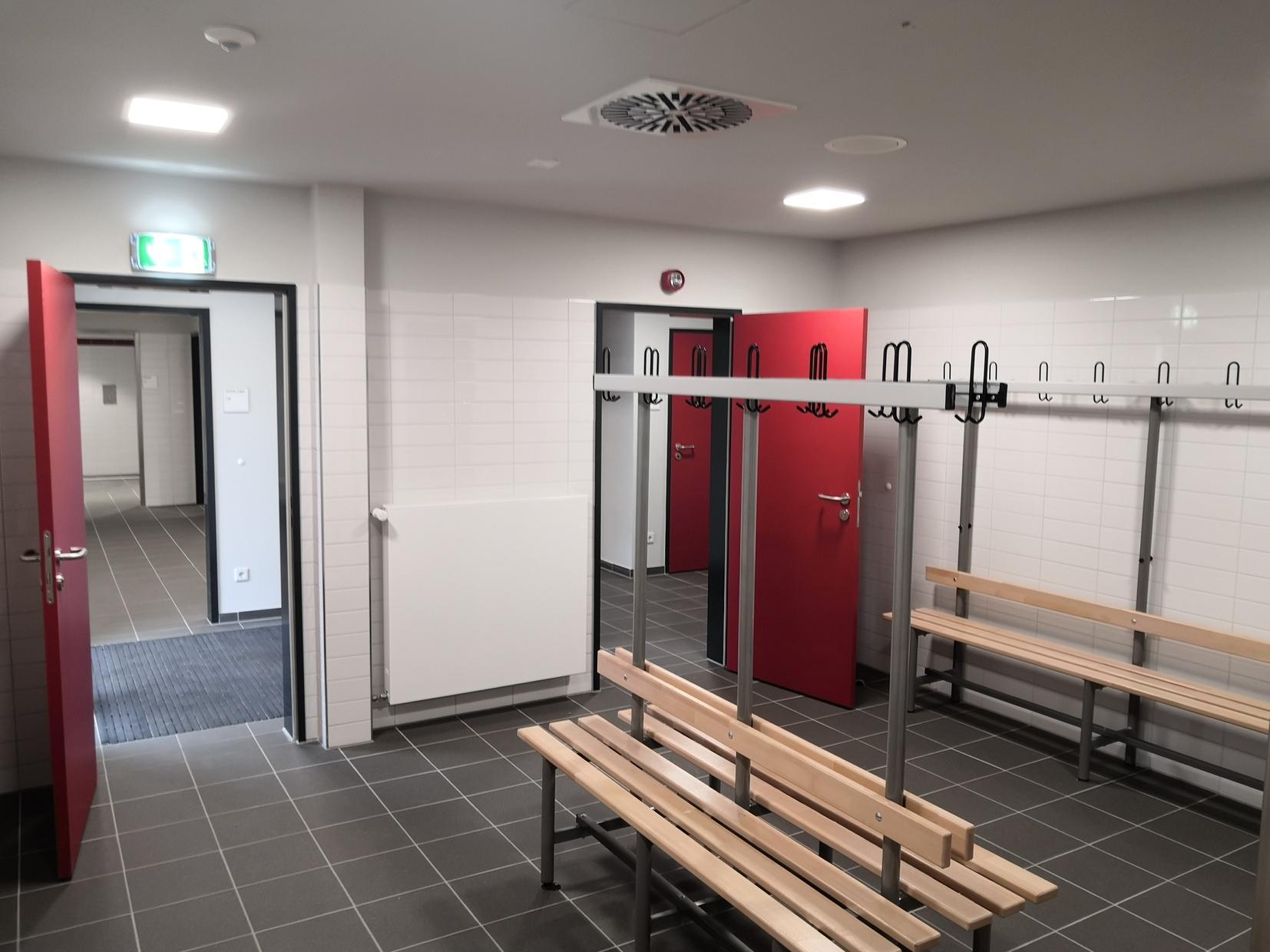Innenansicht eines Umkleideraums mit Holzbank, Kleiderhaken und roten Türen auf weiß gefliestem Boden.