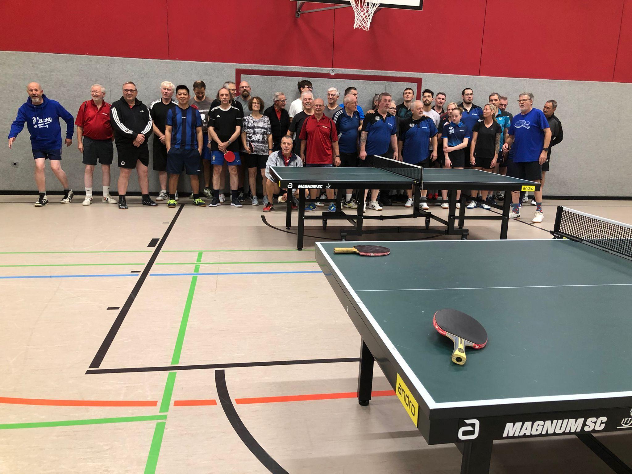 Gruppe von Tischtennisspielern steht hinter zwei Tischtennisplatten in einer Sporthalle, Schläger und Ball auf dem Tisch.