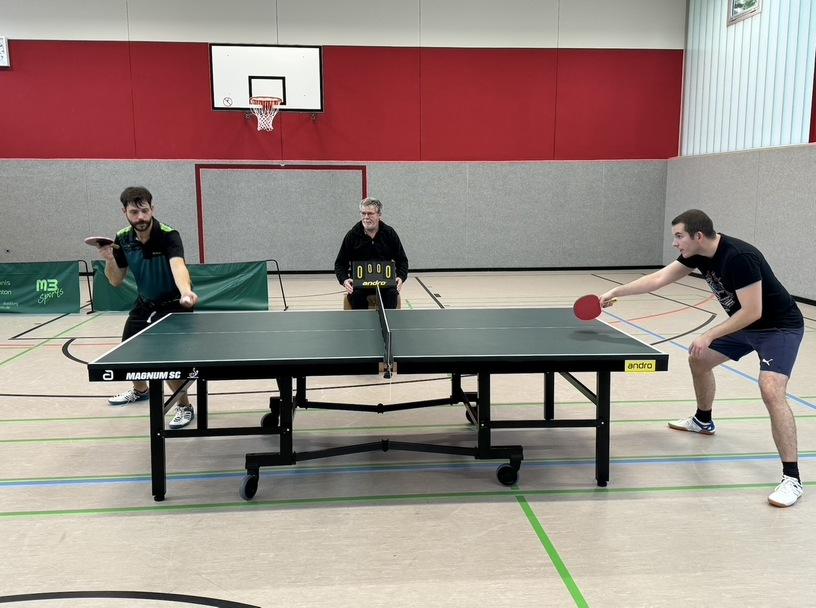Drei M&auml;nner spielen Tischtennis an einem Tisch, einer schl&auml;gt einen Ball, zwei stehen bereit, ein Basketballkorb im Hintergrund.