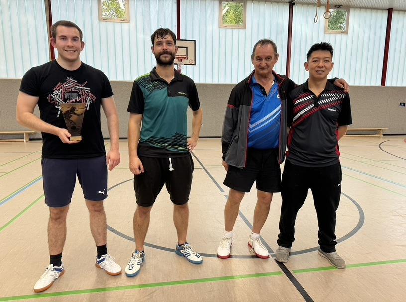 Vier M&auml;nner stehen in Sportkleidung l&auml;chelnd auf einem Tischtennisplatz, Nahaufnahme im Innenbereich.