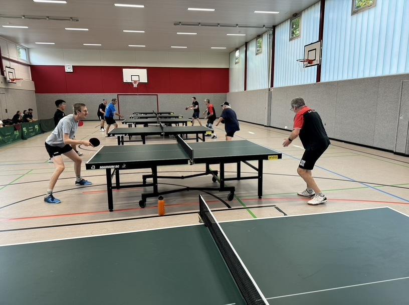 Tischtennisspieler in einem Sportraum spielen an mehreren Tischen mit Schl&auml;gern und B&auml;llen.