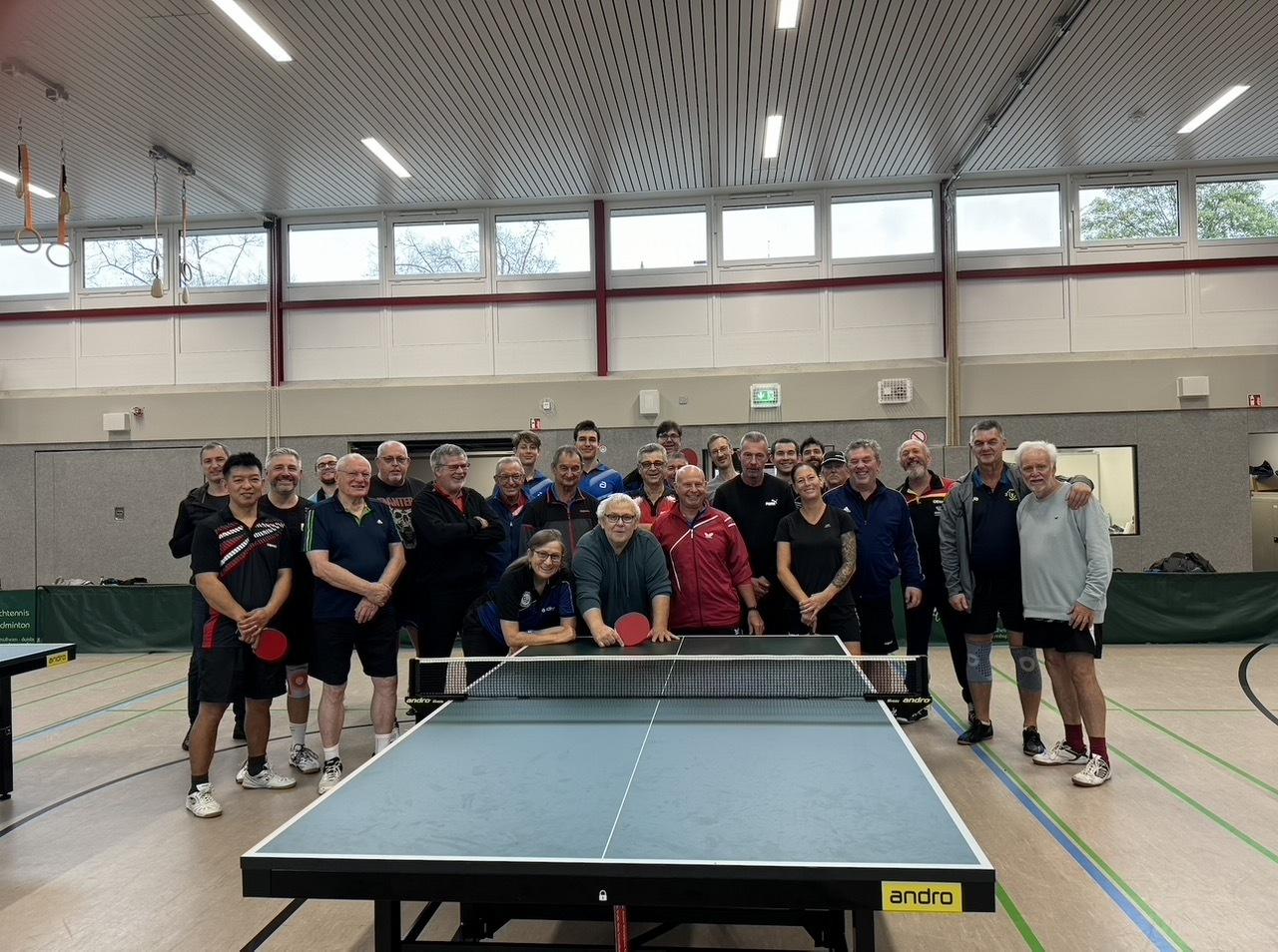 Gruppenfoto von Tischtennisspielern in einer Halle, alle stehen um einen Tischtennistisch herum.