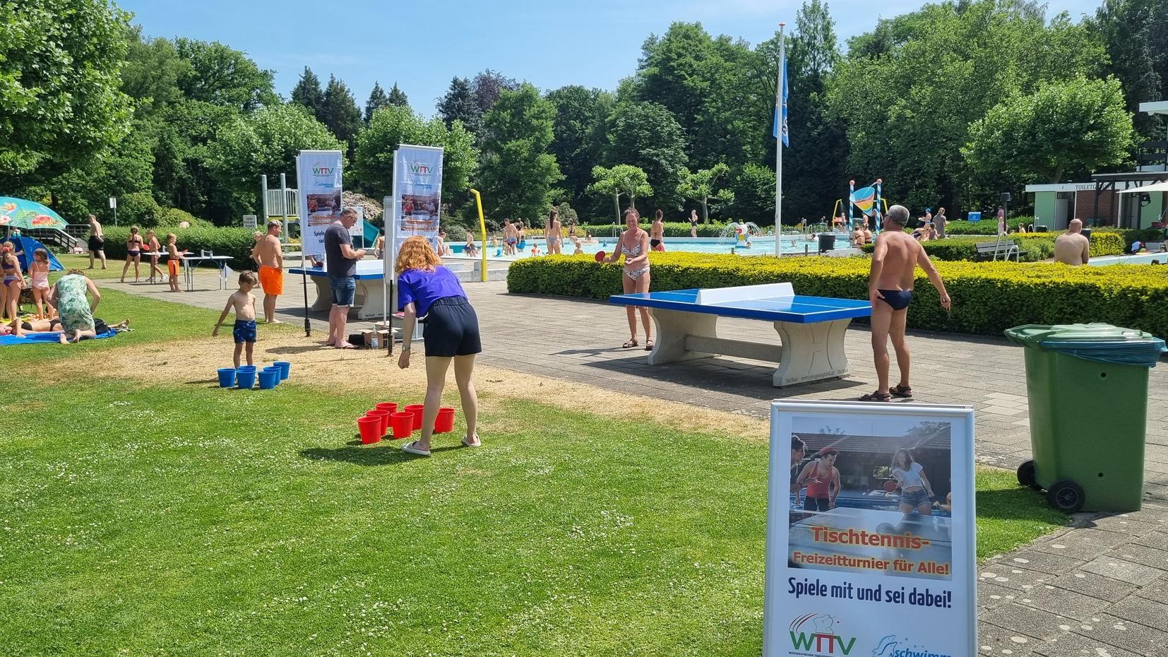 Familien und Kinder spielen im Freibad, neben einer Tischtennisplatte mit Werbeschildern für Tischtennisaktivitäten.
