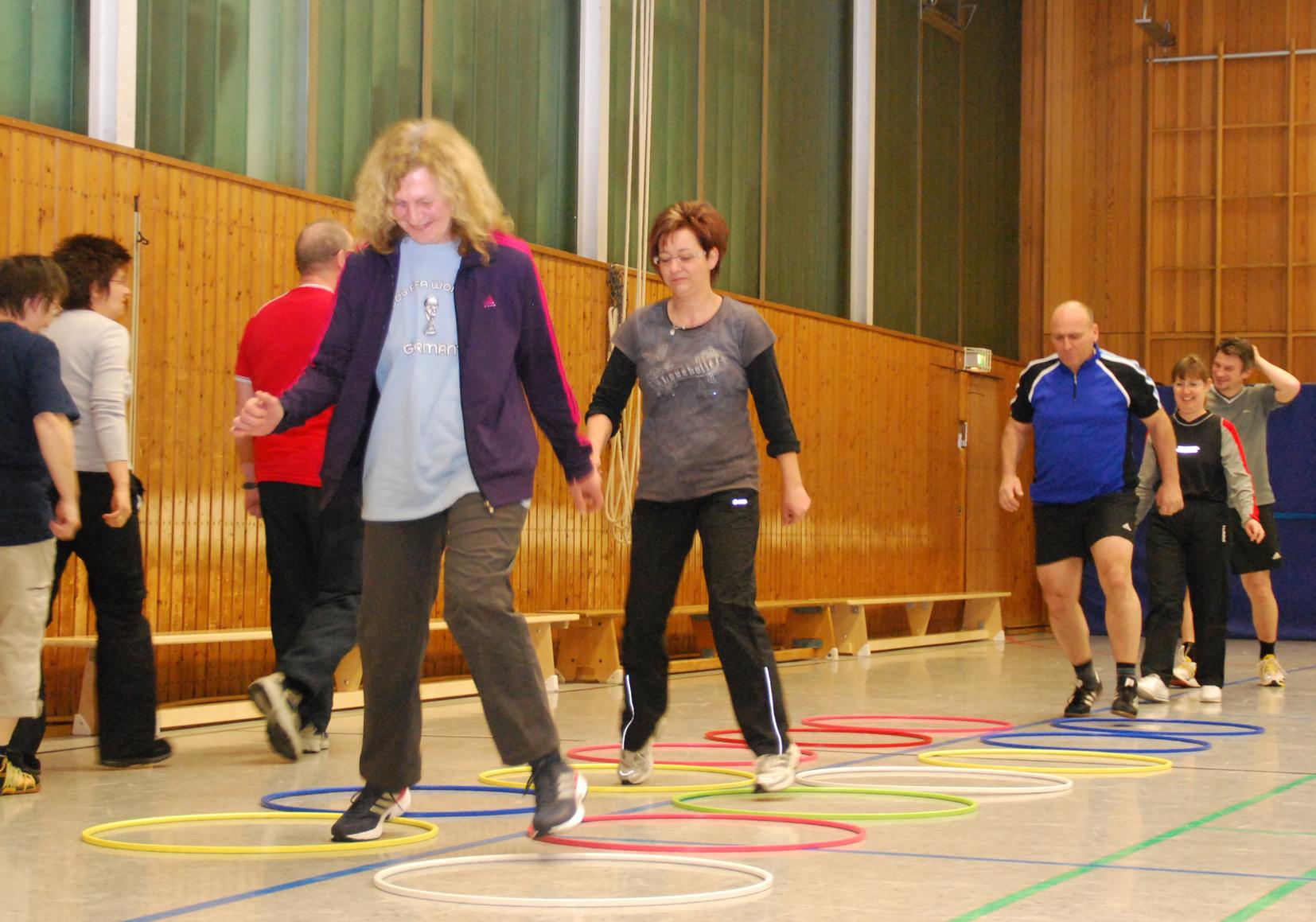 Gruppenteilnehmer bewegen sich in einer Sporthalle von einem Hula-Hoop-Reifen zum anderen.