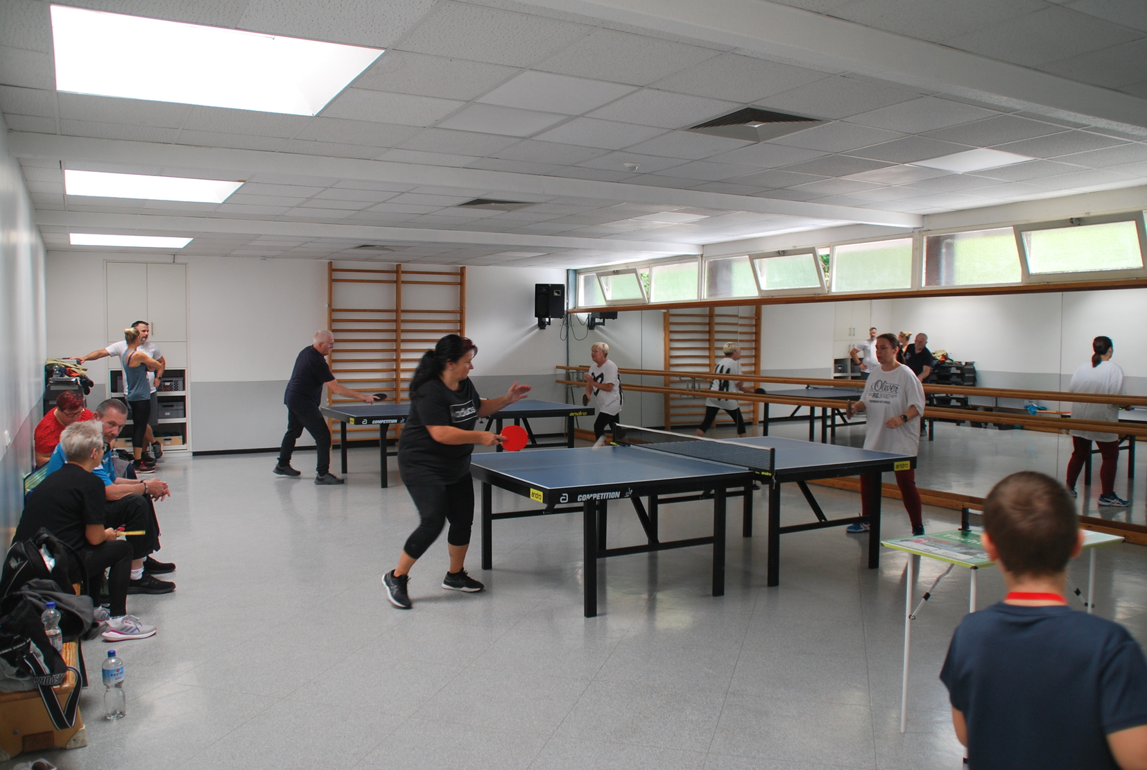Mehrere Personen spielen Tischtennis in einem hellen Raum mit mehreren Tischen und B&auml;nken im Hintergrund.