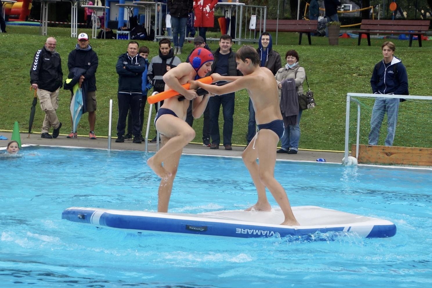 Zwei Jungen k&auml;mpfen auf einem wackeligen Wasser-Trampolin im Schwimmbad, w&auml;hrend Zuschauer am Rand beobachten.