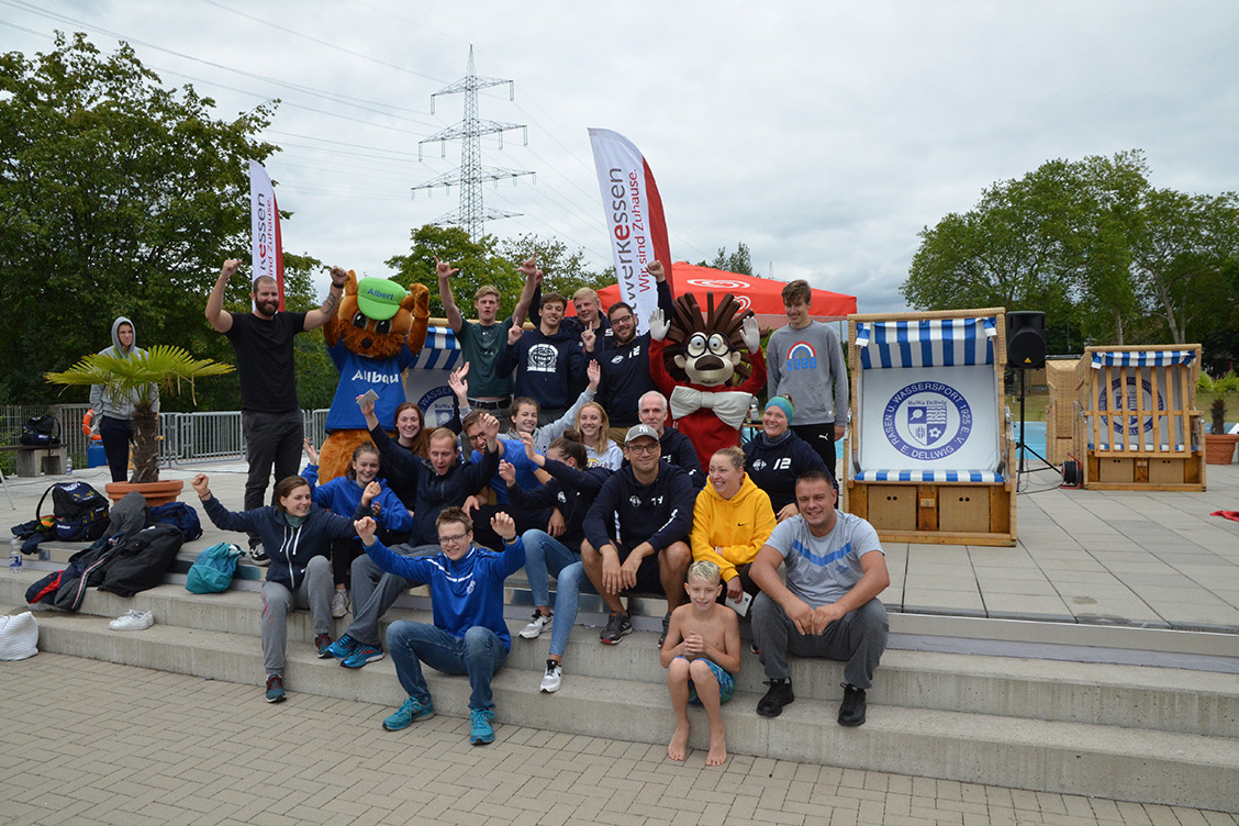 Gruppe von Menschen in sportlichen Outfits posiert fr&ouml;hlich in einem Freiluftbereich mit Bannern und Maskottchen.