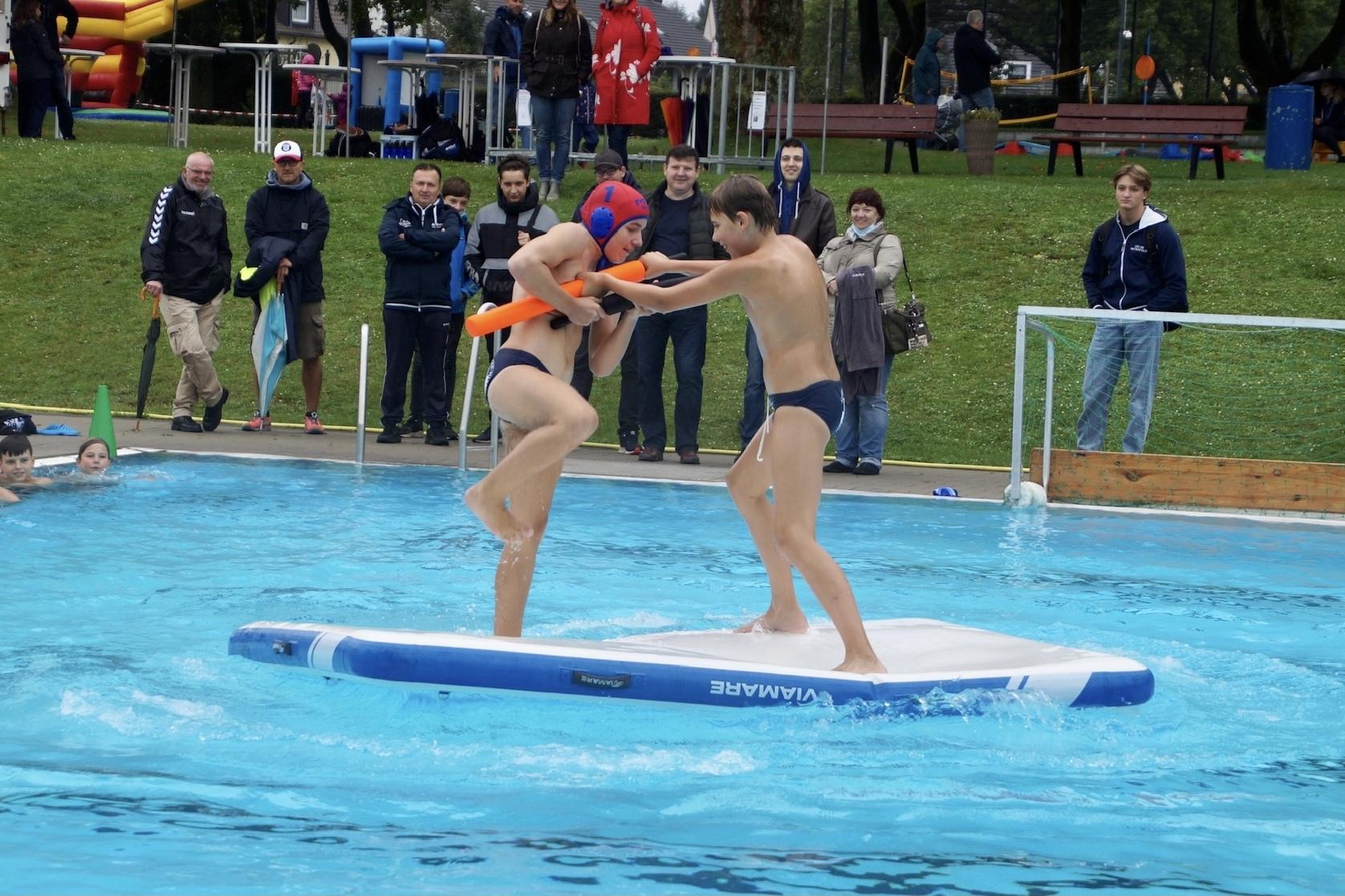 Zwei Jungen k&auml;mpfen auf einem wackeligen Wasser-Trampolin im Schwimmbad, w&auml;hrend Zuschauer am Rand beobachten.
