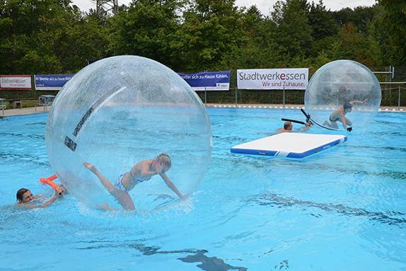 Kinder spielen in Wasserballons im Schwimmbad, w&auml;hrend ein weiteres Kind auf einem Schwimmbrett balanciert.