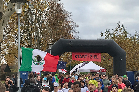 Eine Gruppe von L&auml;ufern passiert das Start-/Zielbanner mit mexikanischer Flagge im Hintergrund. Herbstliche B&auml;ume sichtbar.