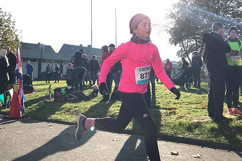 L&auml;uferin in pinkem Oberteil und schwarzer Hose sprintet bei einem Wettlauf, Zuschauer im Hintergrund auf Gr&uuml;nfl&auml;che.