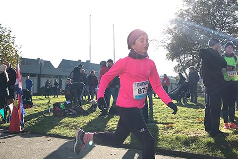 L&auml;uferin in pinkem Oberteil und schwarzer Hose sprintet bei einem Wettlauf, Zuschauer im Hintergrund auf Gr&uuml;nfl&auml;che.