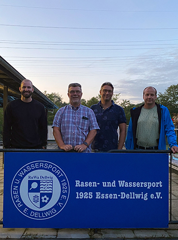 Vier M&auml;nner stehen vor einem blauen Schild mit dem Text &bdquo;Rasen- und Wassersport 1925 Essen-Dellwig e.V.&ldquo;.