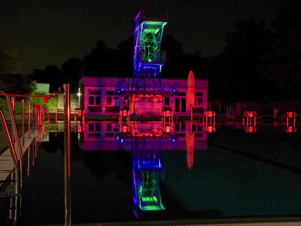 Beleuchteter Schwimmbeckenumrandung mit Sprungturm und reflektierenden Wasseroberfl&auml;che bei Nacht.