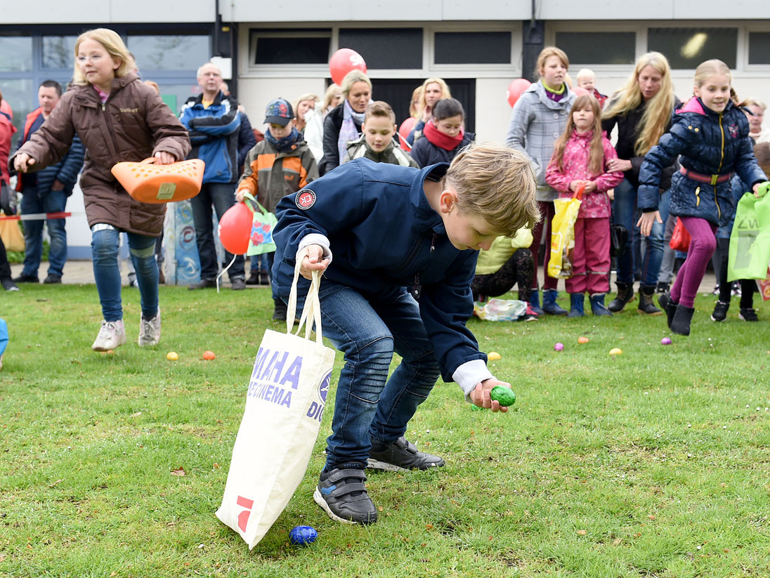 Kind hebt ein buntes Plastik-Ei auf, w&auml;hrend andere Kinder und Erwachsene im Hintergrund auf einer Wiese stehen.