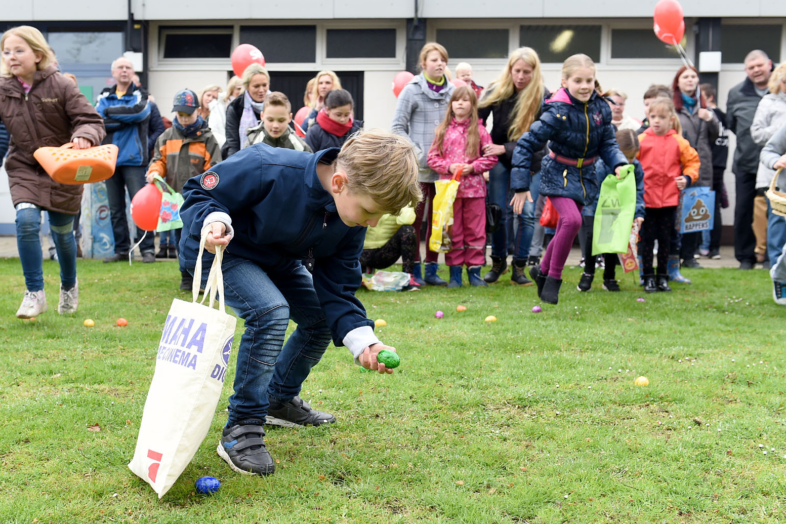 Kind hebt ein buntes Plastik-Ei auf, w&auml;hrend andere Kinder und Erwachsene im Hintergrund auf einer Wiese stehen.