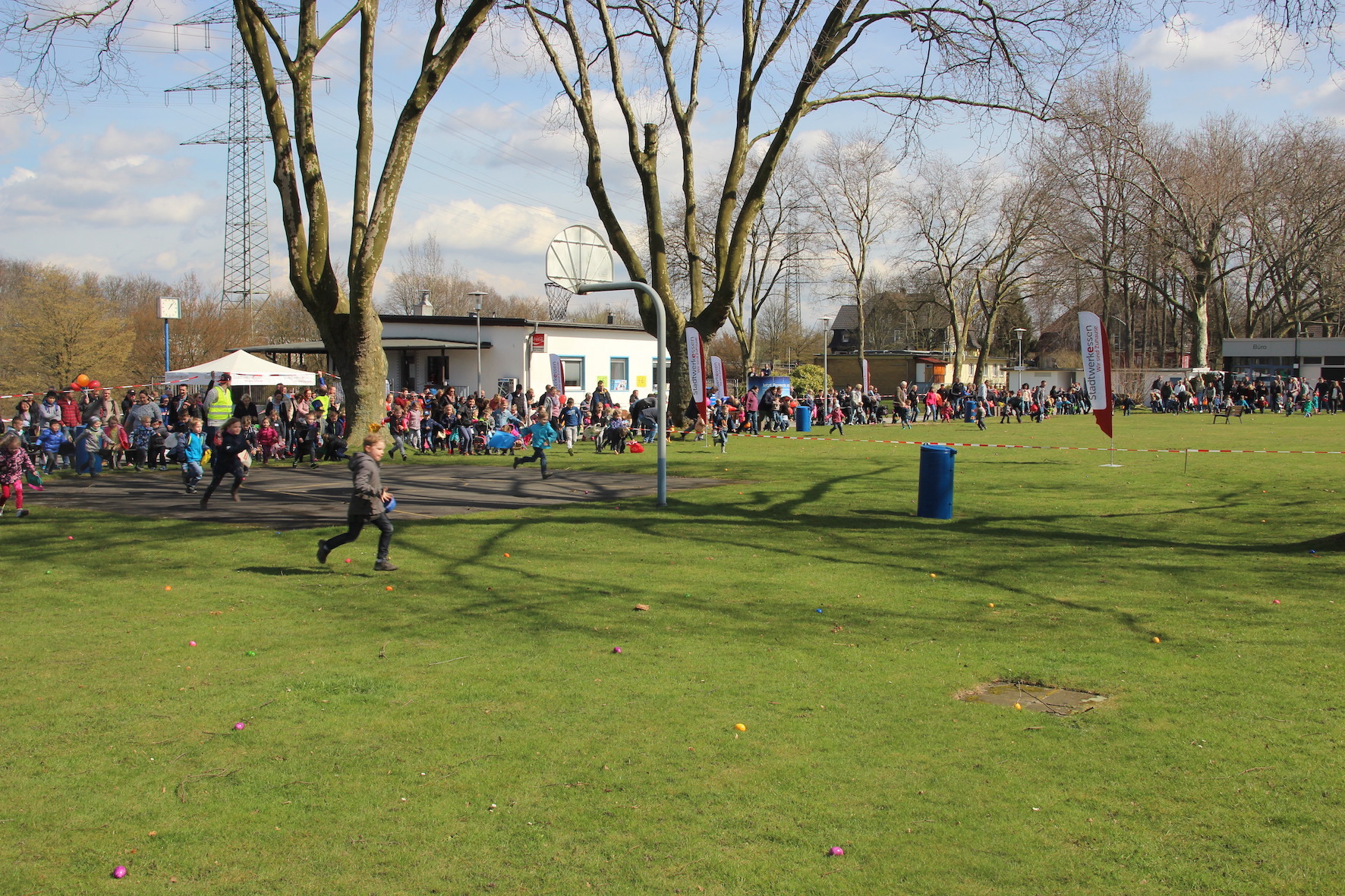 Kinder laufen auf einer Wiese, im Hintergrund ist eine gro&szlig;e Menschenmenge und ein Veranstaltungszelt zu sehen.