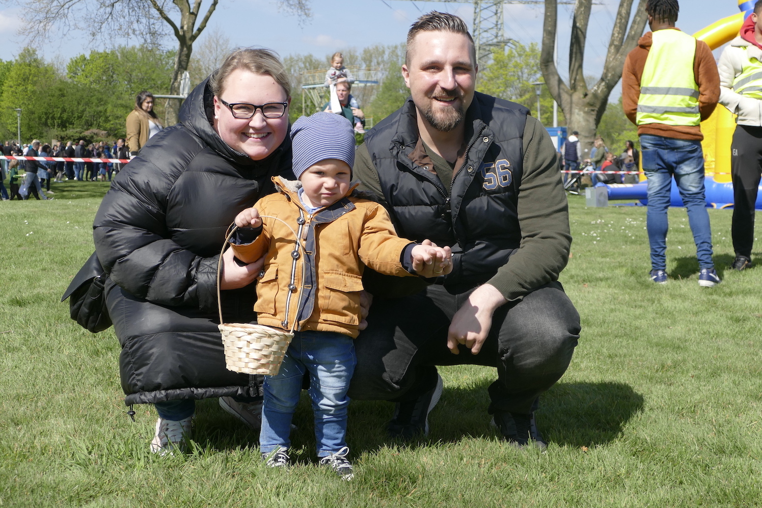 Familie mit einem Kleinkind in der Mitte, das einen Korb h&auml;lt, steht auf einer Wiese an einem sonnigen Tag.